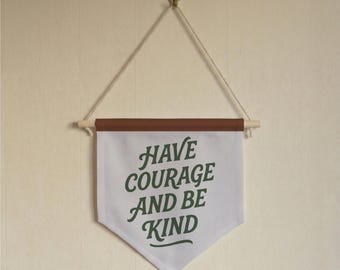 May include: A white banner with the words "HAVE COURAGE AND BE KIND" in green script. The banner has a brown top and is suspended by a wooden dowel and a natural rope. The banner is hanging on a cream-colored wall.