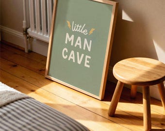 May include: A framed print with the text "little MAN CAVE" in white on a sage green background. The frame is light wood. A small wooden stool with three legs sits nearby on a hardwood floor.