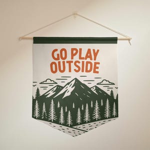 May include: A wall hanging with the text "GO PLAY OUTSIDE" in orange letters. The design features a mountain range and pine trees in green and white, with a light-colored background. The banner is suspended from a wooden dowel.