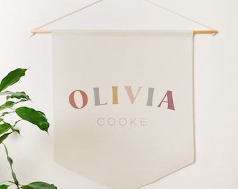 May include: A beige canvas banner with the name "OLIVIA COOKE" printed in colorful, stylized letters. The banner is hanging from a wooden dowel rod with string.