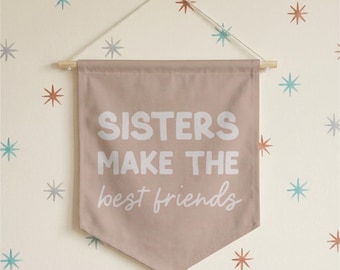 May include: A dusty rose fabric wall banner featuring the white text "SISTERS MAKE THE best friends" in a mix of bold block and cursive lettering. The banner is suspended from a light wooden dowel with a thin white string for hanging.