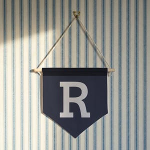 May include: A navy blue pennant banner with the letter "R" in white. The banner is suspended from a wooden dowel and a white string, hanging against a striped blue and white wall.