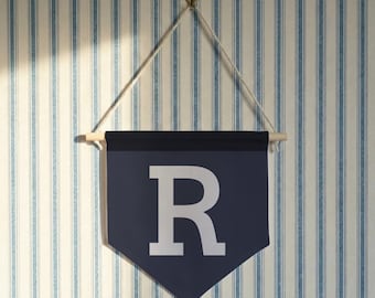 May include: A navy blue pennant banner with the letter "R" in white. The banner is suspended from a wooden dowel and a white string, hanging against a striped blue and white wall.