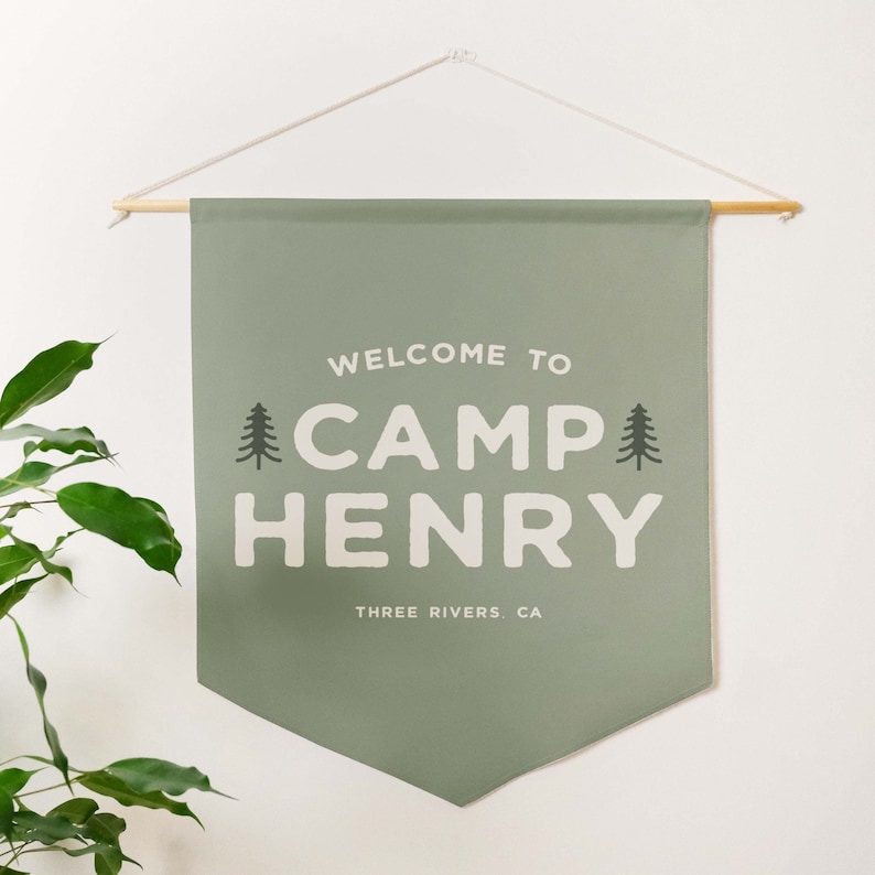 Peut inclure: Une banni&egrave;re murale en tissu vert sauge suspendue &agrave; une tige en bois avec une ficelle blanche. La banni&egrave;re pr&eacute;sente le texte blanc "WELCOME TO CAMP HENRY" avec deux ic&ocirc;nes de sapin vert fonc&eacute; de chaque c&ocirc;t&eacute;, et "THREE RIVERS, CA" en dessous.