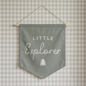 May include: A sage green fabric wall banner featuring the white text Little Explorer in a mix of block and script fonts. A small white pine tree icon is centered below the text. The banner hangs from a wooden dowel with a white string.