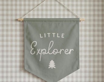 May include: A sage green fabric wall banner featuring the white text Little Explorer in a mix of block and script fonts. A small white pine tree icon is centered below the text. The banner hangs from a wooden dowel with a white string.