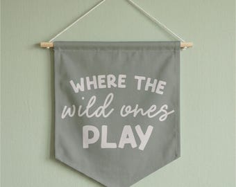 May include: A sage green fabric wall banner featuring the white text WHERE THE wild ones PLAY. The banner is suspended from a light-colored wooden dowel with a white string attached to the ends for hanging, creating a pointed bottom edge.