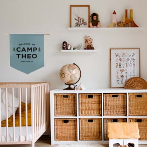 Peut inclure: Une banni&egrave;re murale en tissu bleu suspendue &agrave; une tige en bois avec une ficelle blanche. La banni&egrave;re pr&eacute;sente un texte sombre indiquant WELCOME TO CAMP THEO et THREE RIVERS, CA, encadr&eacute; par deux petites illustrations de sapins verts.