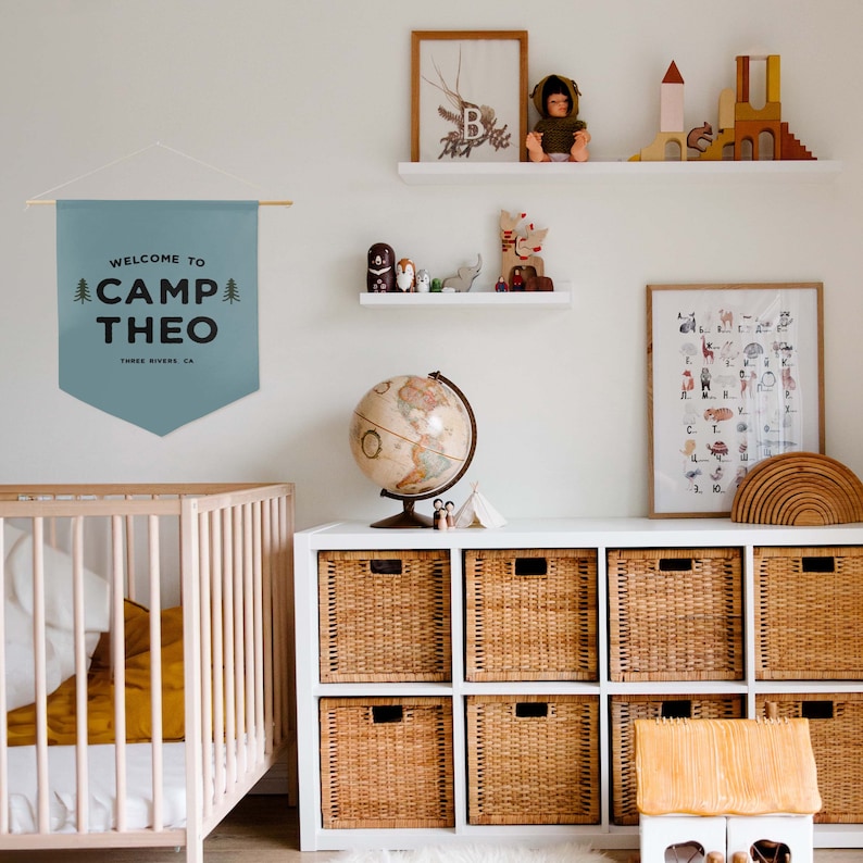 Peut inclure: Une banni&egrave;re murale en tissu bleu suspendue &agrave; une tige en bois avec une ficelle blanche. La banni&egrave;re pr&eacute;sente un texte sombre indiquant WELCOME TO CAMP THEO et THREE RIVERS, CA, encadr&eacute; par deux petites illustrations de sapins verts.