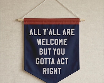 May include: A navy blue fabric wall banner with a pointed bottom edge, featuring the white text ALL Y'ALL ARE WELCOME BUT YOU GOTTA ACT RIGHT. The banner is suspended from a wooden dowel with a white string and has a dark red top border.