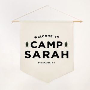 Peut inclure: Une banni&egrave;re murale en tissu cr&egrave;me suspendue &agrave; une tige en bois avec une ficelle blanche. La banni&egrave;re pr&eacute;sente le texte noir "WELCOME TO CAMP SARAH" avec deux petites ic&ocirc;nes de sapin vert de chaque c&ocirc;t&eacute;, et "STILLWATER, OK" imprim&eacute; en dessous.