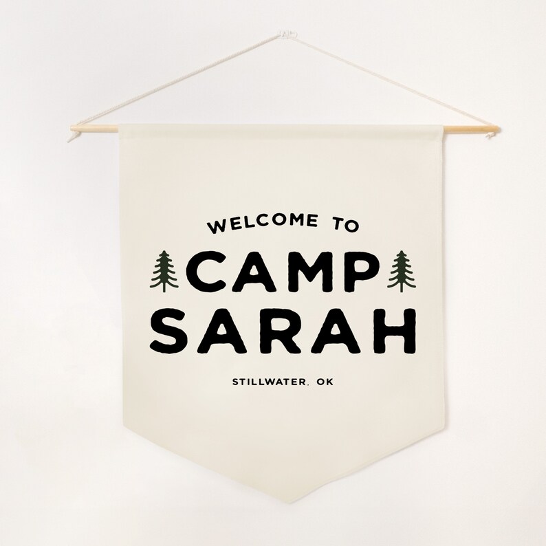 Peut inclure: Une banni&egrave;re murale en tissu cr&egrave;me suspendue &agrave; une tige en bois avec une ficelle blanche. La banni&egrave;re pr&eacute;sente le texte noir "WELCOME TO CAMP SARAH" avec deux petites ic&ocirc;nes de sapin vert de chaque c&ocirc;t&eacute;, et "STILLWATER, OK" imprim&eacute; en dessous.