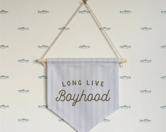 May include: A light blue pennant banner with the text "LONG LIVE Boyhood" in brown script. The banner is suspended by a wooden dowel and a natural rope. The background is a cream-colored wall with a repeating pattern of blue fish.