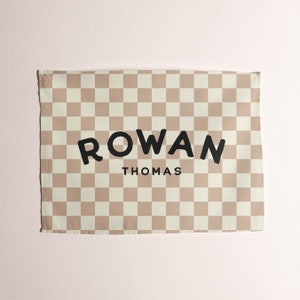 May include: A beige and white checkered fabric banner with the text "ROWAN THOMAS" in black.