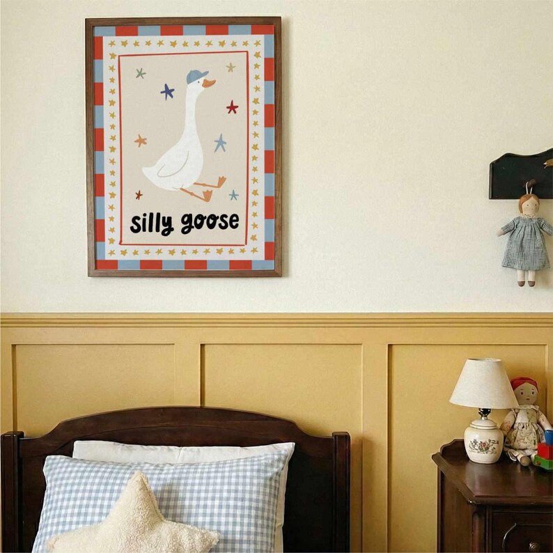 May include: A framed art print featuring a whimsical illustration of a white goose wearing a blue cap. The artwork is bordered with red and blue checkered patterns and includes the text "silly goose". The print is displayed in a child's bedroom.