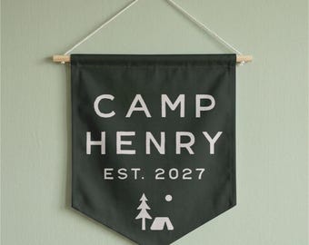 May include: A dark green fabric wall banner featuring the white text CAMP HENRY EST. 2027. Below the text is a simple white graphic of a pine tree, a circle representing the sun or moon, and a tent. The banner hangs from a wooden dowel with a cord.