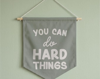 May include: A sage green fabric wall banner featuring the white text "YOU CAN do HARD THINGS" in a mix of block and script fonts. The banner is suspended from a light-colored wooden dowel with a white string attached for hanging on a wall.