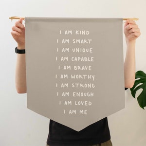 May include: A beige fabric banner with white text that reads "I am kind, I am smart, I am unique, I am capable, I am brave, I am worthy, I am strong, I am enough, I am loved, I am me".