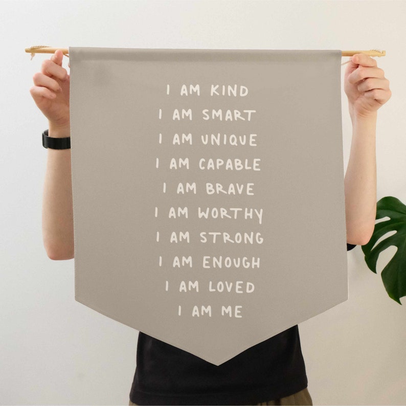 May include: A beige fabric banner with white text that reads "I am kind, I am smart, I am unique, I am capable, I am brave, I am worthy, I am strong, I am enough, I am loved, I am me".