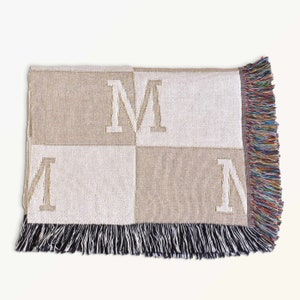 May include: A woven throw blanket with a checkered pattern in beige and white. The blanket features the letter "M" repeated in a larger size in the center and smaller size in the corners. The blanket has black and multicolored fringe.