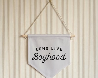 May include: A light gray banner with the words "LONG LIVE Boyhood" in black script. The banner is suspended from a wooden dowel and hung by a string from a gold-colored hook. The background is a striped wall.