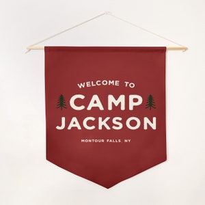Peut inclure: Une banni&egrave;re murale en tissu rouge fonc&eacute; avec le texte blanc WELCOME TO CAMP JACKSON et MONTOUR FALLS, NY. Deux petites ic&ocirc;nes de sapin vert fonc&eacute; entourent le mot CAMP. La banni&egrave;re est suspendue &agrave; une tige en bois avec une ficelle blanche.