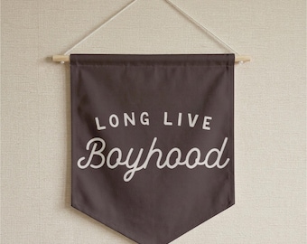 May include: A dark brown fabric wall banner featuring the white text "LONG LIVE Boyhood" in a combination of block and script lettering. The banner is suspended from a wooden dowel with a white string attached for hanging on a wall.