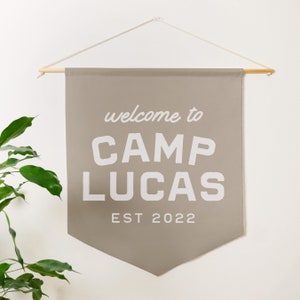 May include: A beige canvas banner with the text "welcome to CAMP LUCAS EST 2022" in white. The banner is hanging from a wooden dowel rod with white string.