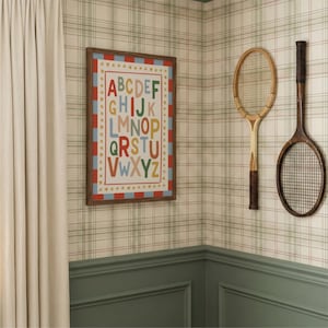May include: A framed alphabet print with colorful letters hangs on a plaid wallpapered wall. Two vintage wooden tennis rackets are mounted nearby. The room features a green wainscoting and a cream-colored curtain.