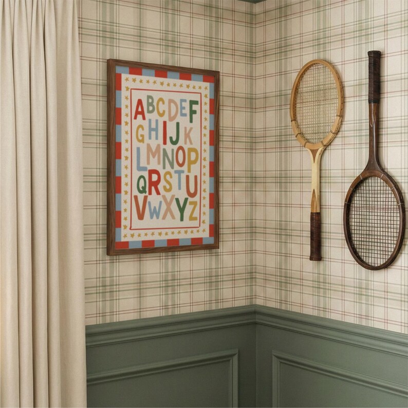 May include: A framed alphabet print with colorful letters hangs on a plaid wallpapered wall. Two vintage wooden tennis rackets are mounted nearby. The room features a green wainscoting and a cream-colored curtain.