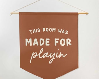 May include: A terracotta-colored banner with the text "THIS ROOM WAS MADE FOR playin" in white. The banner is suspended from a wooden dowel with a white cord for hanging. The banner has a pointed bottom.