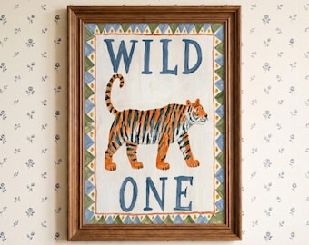 May include: A framed wall art print featuring a hand-painted style tiger in orange and black stripes. The text WILD ONE is written in blue above and below the tiger. The border consists of a repeating pattern of green and blue triangles.