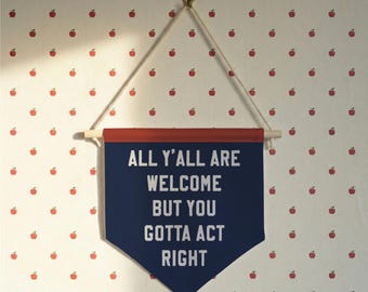 May include: A navy blue banner with the text "ALL Y'ALL ARE WELCOME BUT YOU GOTTA ACT RIGHT" in white lettering. The banner has a red top and is suspended by a wooden dowel and string. The background is a white wall with a pattern of red apples.