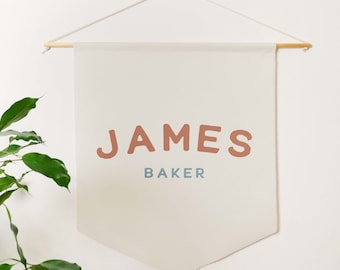 May include: A white canvas banner with the name "JAMES BAKER" printed in brown and blue lettering. The banner is hanging from a wooden dowel rod with string.