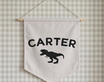 May include: A cream-colored fabric wall banner featuring the name CARTER printed in bold black capital letters above a black silhouette of a Tyrannosaurus Rex. The banner is suspended from a light-colored wooden dowel with a white string.
