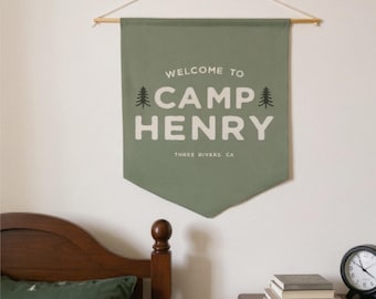 May include: A sage green fabric wall banner featuring the white text WELCOME TO CAMP HENRY and THREE RIVERS CA, flanked by two small black pine tree icons. The banner is suspended from a wooden dowel with a thin string for hanging.