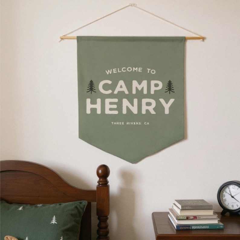 Peut inclure: Une banni&egrave;re murale en tissu vert sauge avec le texte blanc WELCOME TO CAMP HENRY et THREE RIVERS CA, encadr&eacute; par deux petites ic&ocirc;nes de sapins noirs. La banni&egrave;re est suspendue &agrave; une tige en bois avec une fine ficelle pour l'accrocher.