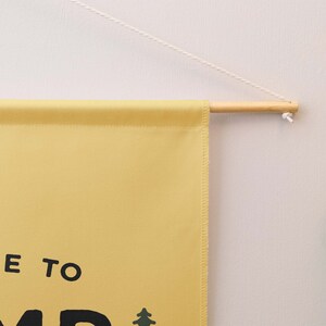 Peut inclure: Une banni&egrave;re murale en tissu jaune avec le texte noir "E TO" et une petite ic&ocirc;ne de sapin vert. La banni&egrave;re est suspendue &agrave; une tige en bois clair par une fine cordelette blanche, formant un affichage simple pour une surface murale.