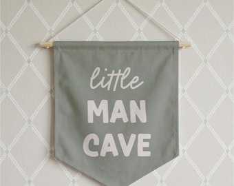 May include: A sage green fabric wall banner featuring the white text "little MAN CAVE" in a mix of script and block lettering. The banner is suspended from a light-colored wooden dowel with a thin white string attached for hanging purposes.