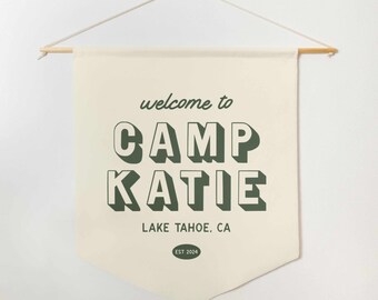 May include: A beige canvas banner with the text "welcome to CAMP KATIE LAKE TAHOE, CA EST 2024" in green lettering. The text is in a retro, 3D style.