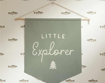 May include: A sage green banner with the words "LITTLE Explorer" in white script and a tree graphic. The banner is suspended from a wooden dowel and string. The background is a white wall with a repeating pattern of fish.