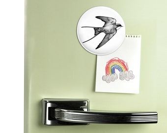 May include: A green refrigerator with a white round magnet featuring a black and white drawing of a bird in flight. A white piece of paper with a rainbow and clouds drawing is attached to the refrigerator with a magnet.
