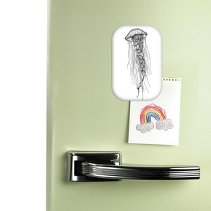 Peut inclure: Un aimant rectangulaire blanc avec une illustration en noir et blanc d'une méduse est fixé sur un réfrigérateur vert clair. Un petit dessin d'un arc-en-ciel est également attaché.