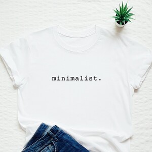 May include: White t-shirt with the text "minimalist." printed in black on the front. The shirt is laid flat on a white surface with a pair of blue jeans and a small potted plant.
