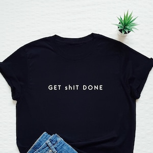 Puede incluir: Camiseta negra con la frase blanca "GET sh!t DONE", combinada con vaqueros azules. Una pequeña planta verde es visible en el fondo. La camiseta está doblada y los vaqueros están cuidadosamente colocados.