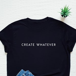 Może przedstawiać: Czarny T-shirt z napisem "CREATE WHATEVER" w białych literach. Koszulka leży na białym tle z niebieskimi dżinsami i małą doniczką z rośliną.