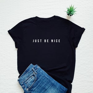 May include: Black t-shirt with white text that reads "JUST BE NICE". The shirt is lying on a white surface with a pair of blue jeans and a small potted plant.