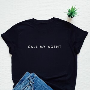 May include: Black t-shirt with white text that reads "CALL MY AGENT". The shirt is laying on a white surface with a pair of blue jeans.