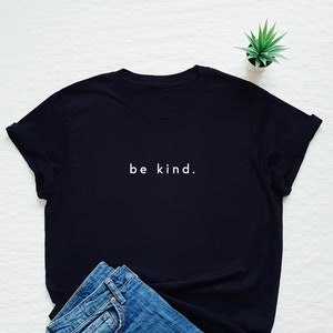 Pode incluir: Camiseta preta com o texto "be kind." em letras brancas. A camiseta está sobre uma superfície branca com um par de calças de ganga azuis e uma pequena planta em vaso.