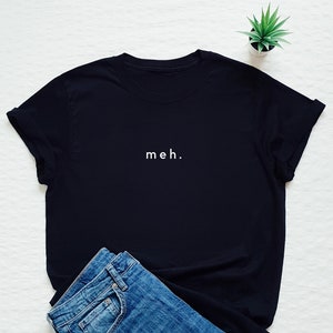 May include: Black t-shirt with the word "meh." printed in white on the front. The shirt is lying on a white surface with a pair of blue jeans folded next to it.